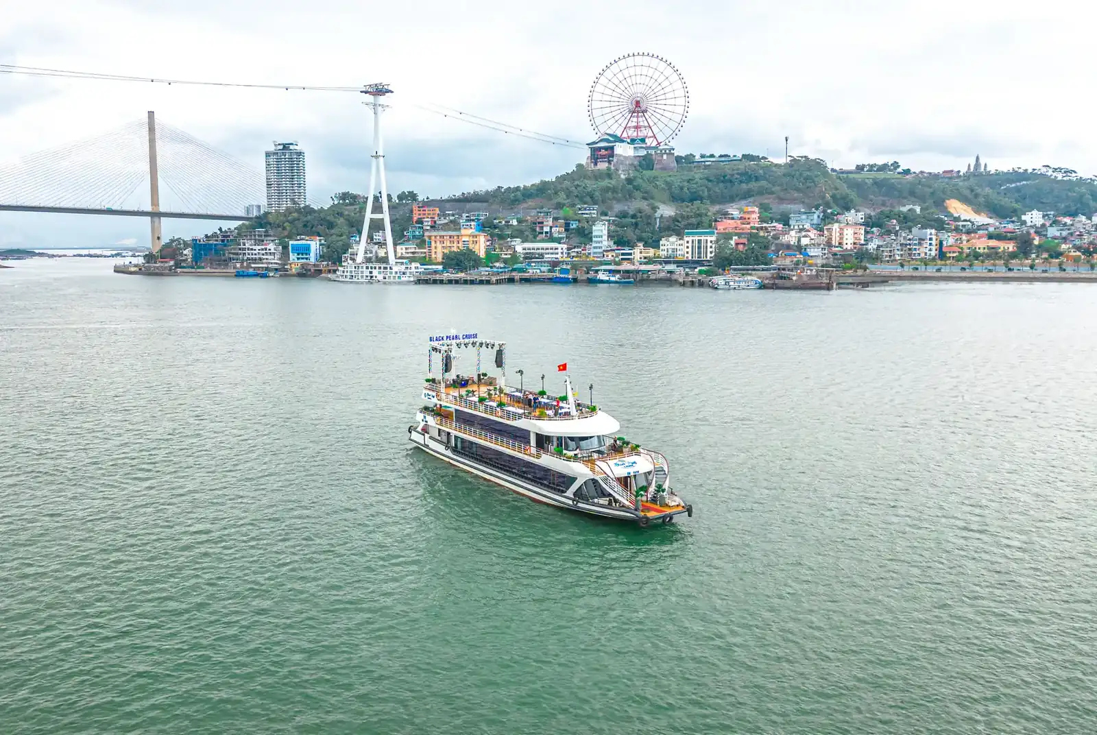 Du thuyền Black Pearl Cruise di chuyển trên vịnh, phía xa là cầu Bãi Cháy và vòng quay Mặt Trời Hạ Long